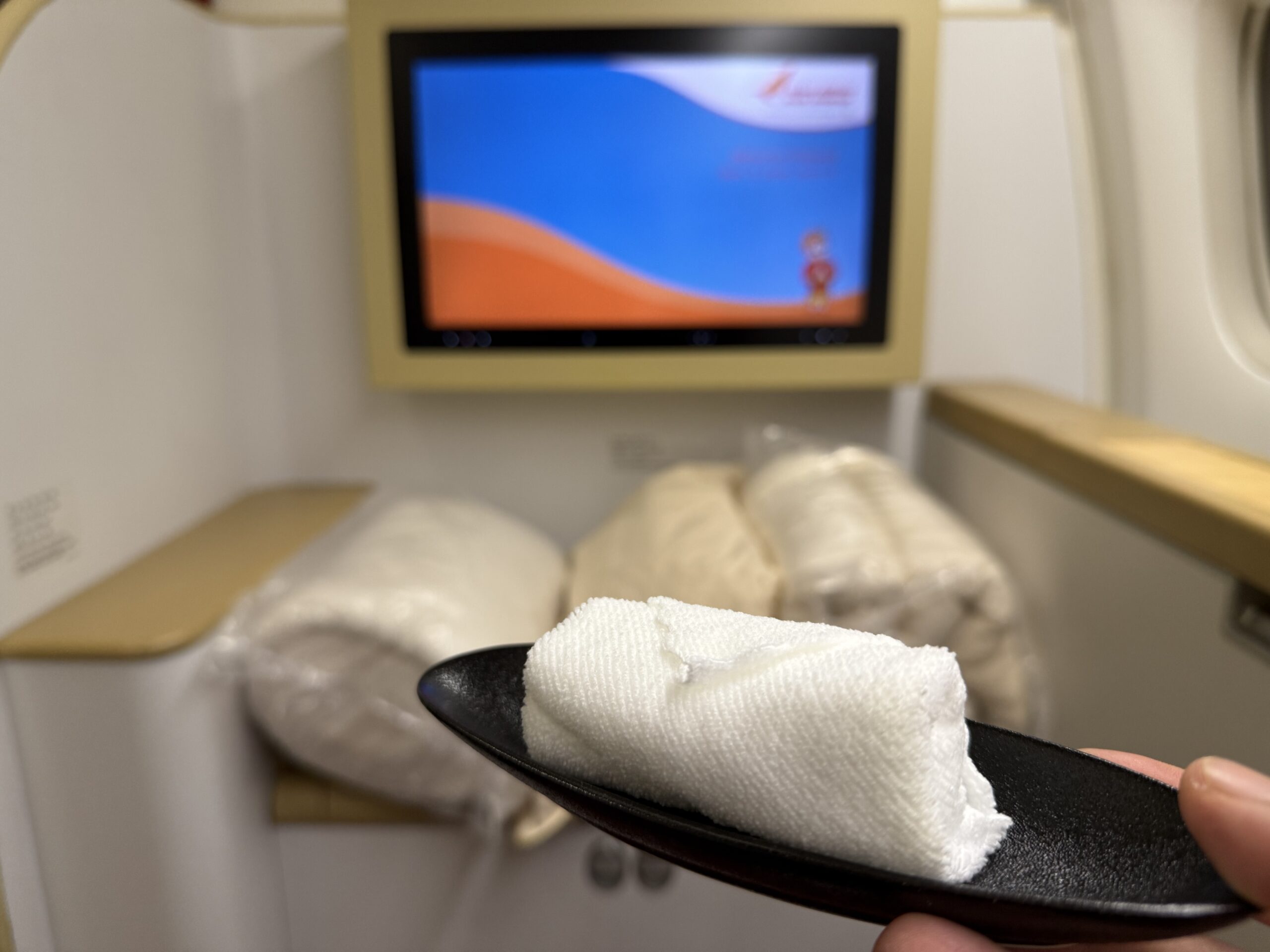 Air India First Class a hand holding a towel on a spoon in front of a television